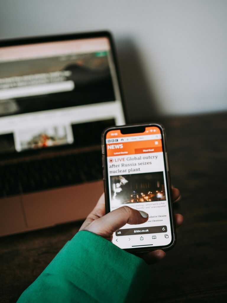 Person using smartphone for reading news online with laptop in background.